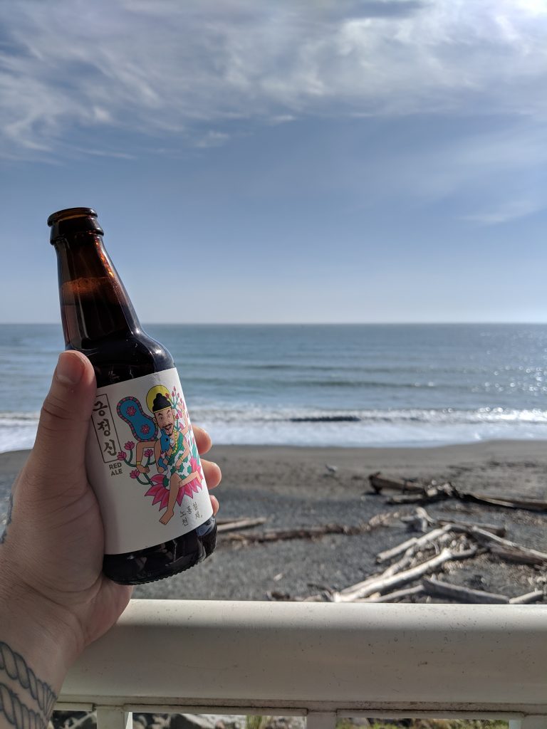 Hand holding a bottle of The Booth red ale against a deck rail, overlooking a southern Oregon coast beach.