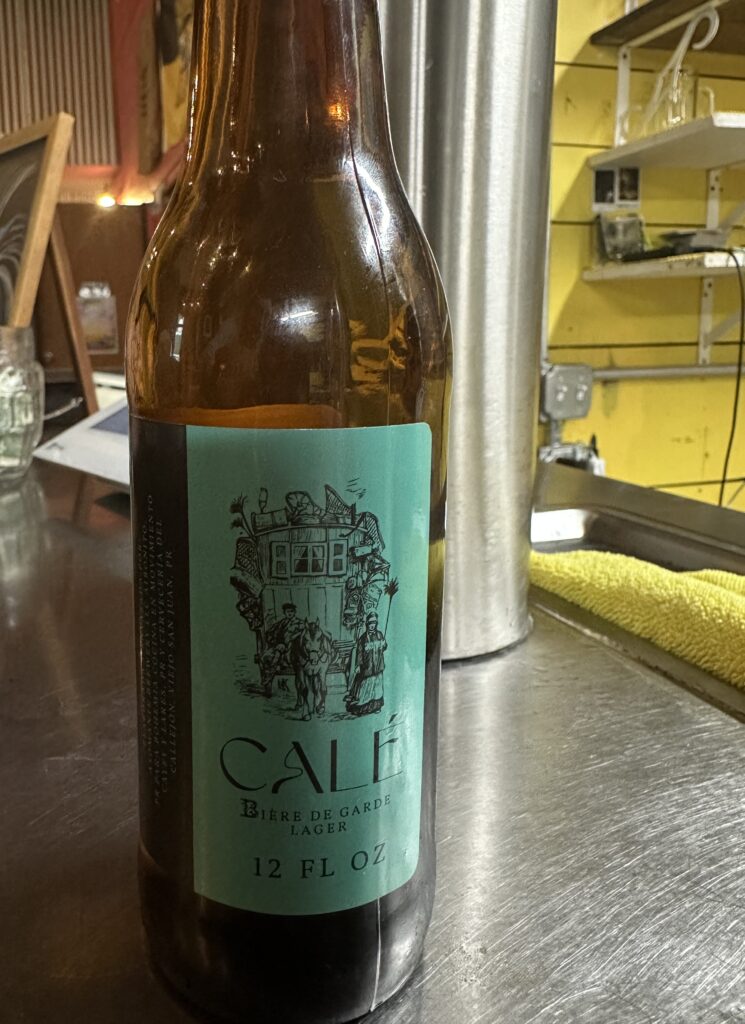 12 ounce brown glass beer bottle showing the label for Calé. The label is teal and says the beer is a Biere de Garde lager. The bottle is sitting on the bar at Cervecería del Callejón.