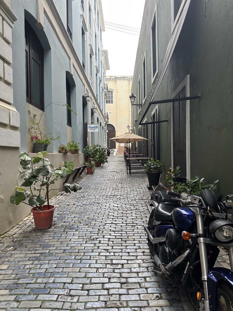 View up Callejón de la Capilla. It's a blue cobblestone alley between Spanish colonial-style buildings. Potted plants line the alley. A parked motorcycle is in the foreground. A small sign for Cervecería del Callejón can be seen further up the street, across from tables and benches with umbrellas shading them.