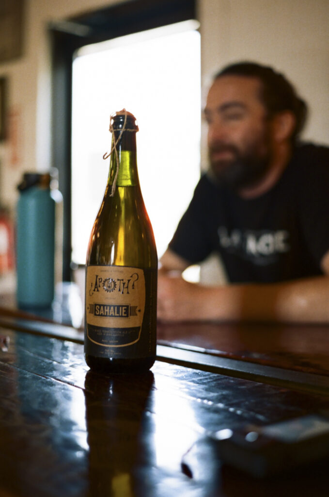 A bottle of Sahalie, a 10.11% spontaneously fermented beer, in The Ale Apothecary’s Bend location, with Connor Currie, brewer, in the background, April 18th, 2026.