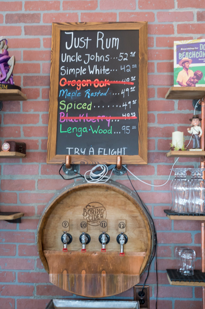 Boring Cider Company barrel sticking out a brick wall, with four taps in the barrel. There's a blackboard menu on the wall above the barrel.