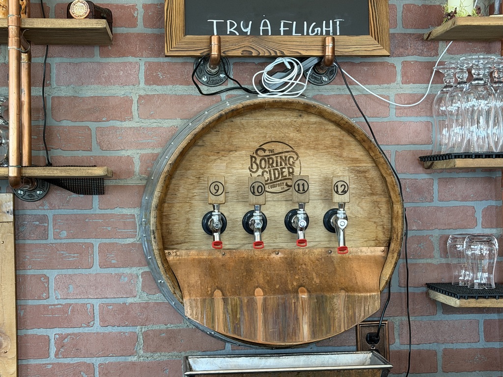 The Boring Cider Company barrel sticking out of a brick wall. There are four taps in the barrel. The bottom of a blackboard can be seen on the wall above the barrel. The blackboard reads, "Try a flight".