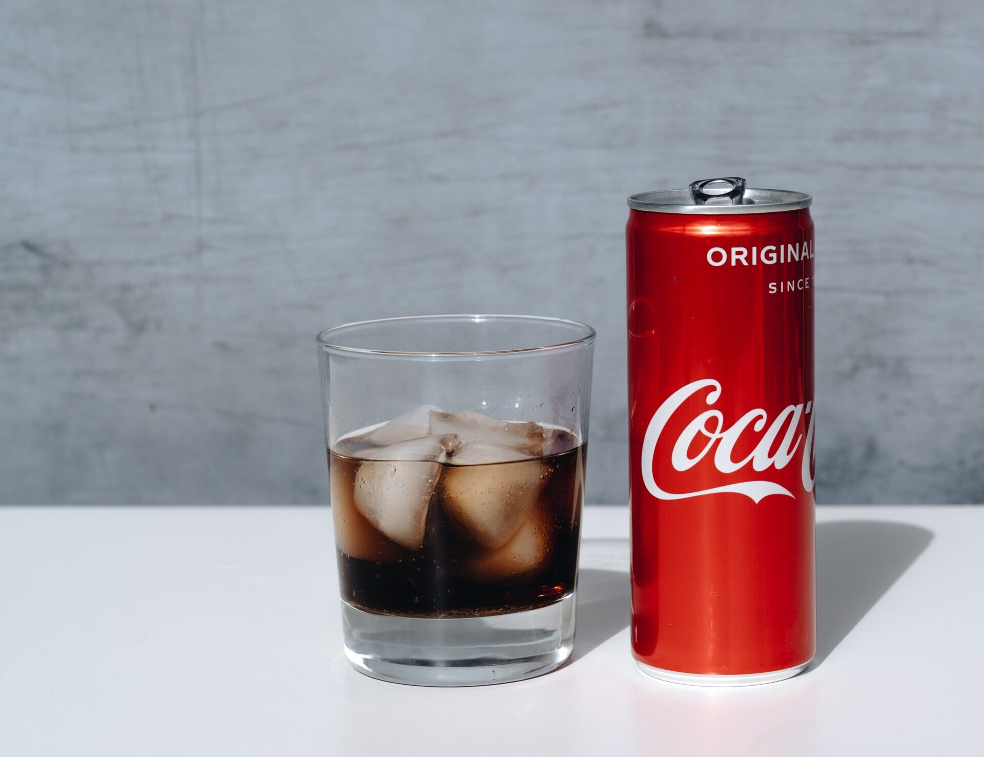 Chilled Coca-Cola can and drink with ice cubes in glass on minimalistic surface.